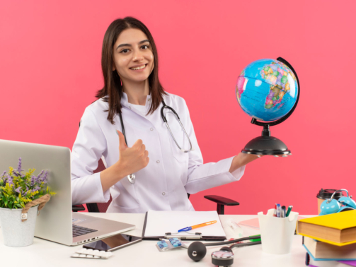young-female-doctor-white-coat-with-stethoscope-around-her-neck-holding-globe-looking-front-with-smile-face-showing-thumbs-up-sitting-table-with-laptop-pink-wall